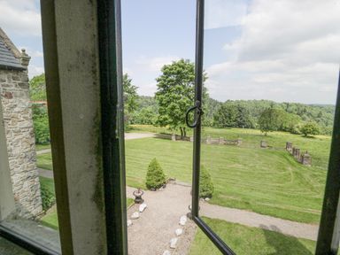View through an open window showing a stone building, a gravel path, trimmed bushes, mowed lawn, and trees at Pentre Hall in Bronygarth near Chirk