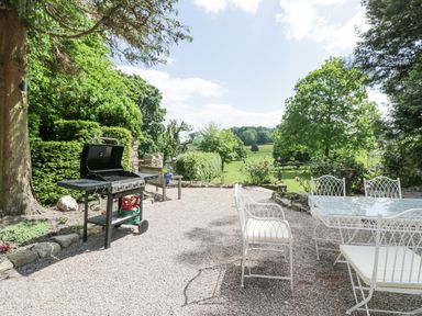 An outdoor patio with a barbecue grill and white metal table and chairs surrounded by trees and greenery at Pentre Hall in Bronygarth near Chirk