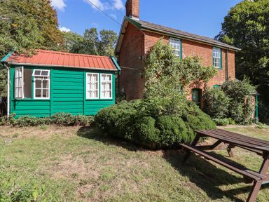 A brick house with green wooden extension and a wooden picnic table in a garden at Warren Cottage in Bures near Assington