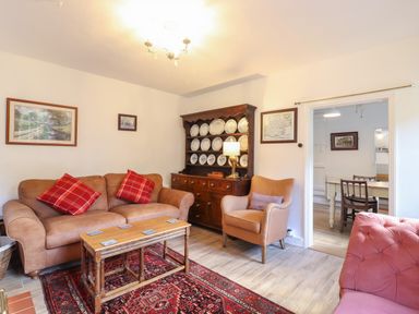 A living room with a brown sofa with red cushions a wooden coffee table and a cabinet with plates at Warren Cottage Bures near Assington