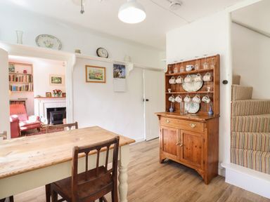 A dining area with a wooden table and chairs a wooden cupboard with hanging mugs and glasses and stairs with striped carpet at Warren Cottage in Bures near Assington