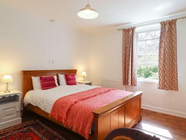 A bedroom with a wooden bed and red patterned bedding next to a window with floral curtains at Warren Cottage in Bures near Assington