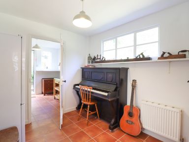 A room with a black upright piano and wooden chair next to an acoustic guitar and a white radiator at Warren Cottage in Bures near Assington