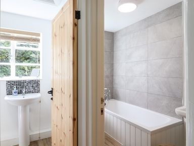 A bathroom with a white bathtub and gray tiled wall and a basin with a window and wooden door at Warren Cottage in Bures near Assington