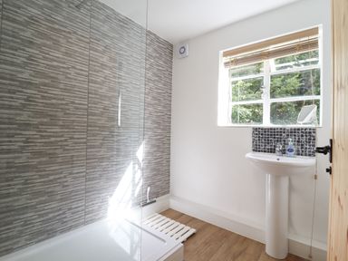 A bathroom with a walk-in shower, a white pedestal sink, a window with blinds, and wooden flooring at Warren Cottage in Bures near Assington