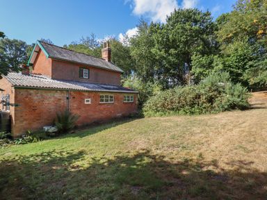 A brick house with a tiled roof next to a grassy yard and surrounded by trees at Warren Cottage in Bures near Assington