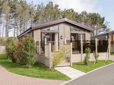A small beige cabin with large windows and glass railing on a green lawn with shrubs at Rockpool Retreat in Runswick Bay near Hinderwell