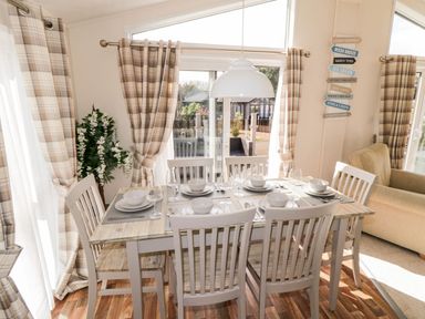 A dining area with a wooden table set for six with white dishes and glassware next to large windows with plaid curtains at Rockpool Retreat in Runswick Bay near Hinderwell