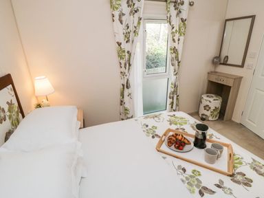 A bedroom with a bed with floral bedding a bedside lamp a dressing table with a mirror and a window with floral curtains at Rockpool Retreat in Runswick Bay near Hinderwell