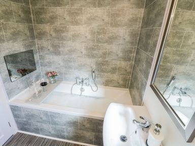 A bathroom with a bathtub partially filled with foam a sink and a mirror with flowers and a glass of wine on the tub at Rockpool Retreat in Runswick Bay near Hinderwell