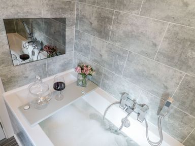 A bathroom with a bathtub filled with bubbles a glass of red wine flowers in a vase and a container labeled bath bombs on a tray across the tub at Rockpool Retreat in Runswick Bay near Hinderwell
