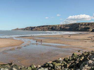 A beach with people walking on the sand and water with cliffs in the background at Rockpool Retreat in Runswick Bay near Hinderwell