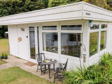 A white exterior corner of a house with large windows a glass door a small round metal table and two chairs a bird feeder and greenery at Ty Haf in Roch