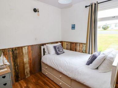 A bedroom with a single bed with white bedding and nautical pillows a wooden floor and a window with striped curtains at Ty Haf in Roch