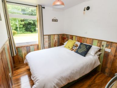 A bedroom with a double bed white bedding cushions and a teddy bear wood paneling on walls and a window with curtains at Ty Haf in Roch