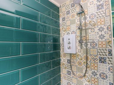 A shower area with a mounted showerhead and control unit on patterned tiles and green rectangular wall tiles at Ty Haf in Roch