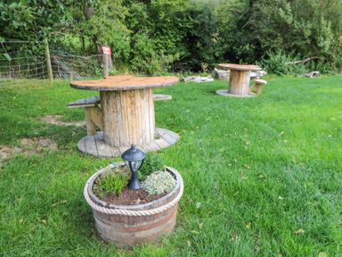 An outdoor area with two wooden spool tables and benches on grass with a planter containing plants and a small lantern at The Salmon in Nantmel near Rhayader