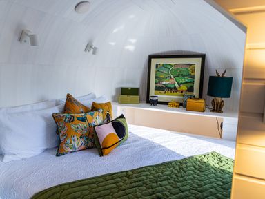A bed with patterned pillows and a green quilt with artwork and decorative items on a shelf in a white curved room at Veiko - Mayflower Meadow in Burshill near Brandesburton