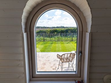 A garden with green grass wooden fence and a chair viewed through an arched window at Veiko - Mayflower Meadow in Burshill near Brandesburton