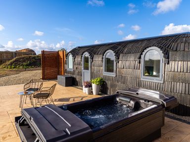 An outdoor patio with a hot tub two chairs and a small table next to a building with arched windows at Veiko - Mayflower Meadow in Burshill near Brandesburton