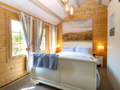 A bedroom with a white bed frame and blue blanket with wooden walls and light curtains at Willow Lodge in Potter Heigham