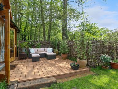 An outdoor wooden deck with a wicker sofa set and potted plants surrounded by trees at Willow Lodge in Potter Heigham