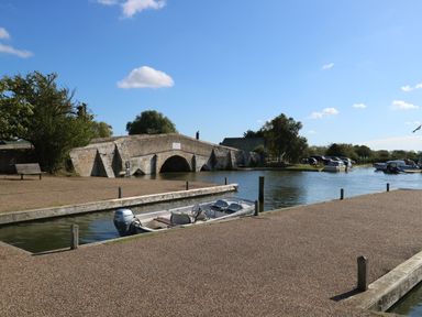 A river with a stone arch bridge and boats docked at a marina at Willow Lodge in Potter Heigham