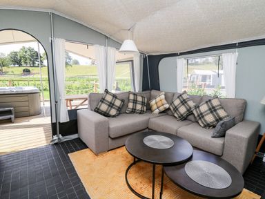 An interior view of a tent with a grey L-shaped sofa with plaid cushions a round black coffee table on a yellow rug and a hot tub outside at The Otter in Nantmel near Rhayader