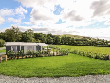 A grassy area with a large tent structure and small shrubs near a fenced field with hills and trees in the background at The Otter in Nantmel near Rhayader