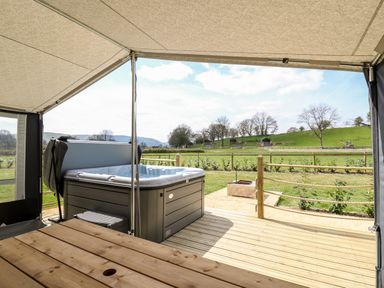 A hot tub on a wooden deck with a picnic table under a canopy overlooking a grassy fenced field at The Otter in Nantmel near Rhayader