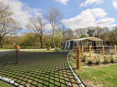 A large net hammock stretched between wooden posts near a pitched tent with a fenced patio and seating area surrounded by trees at The Otter in Nantmel near Rhayader