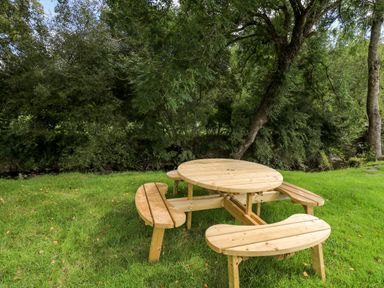 A wooden picnic table with curved benches on grass near trees and a small stream at The Otter in Nantmel near Rhayader