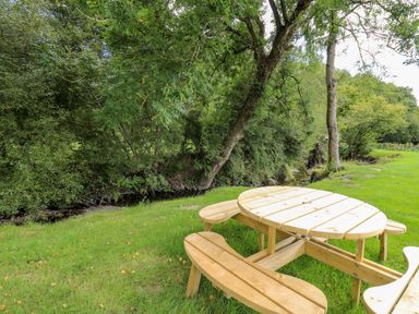 A wooden picnic table with attached benches on grass near trees and a small stream at The Otter in Nantmel near Rhayader