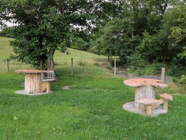 A grassy outdoor area with two wooden cable spool tables converted into seating near a fence and trees at The Otter in Nantmel near Rhayader