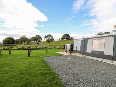 A caravan on a gravel area next to a grassy field with wooden posts and trees in the background at The Otter in Nantmel near Rhayader