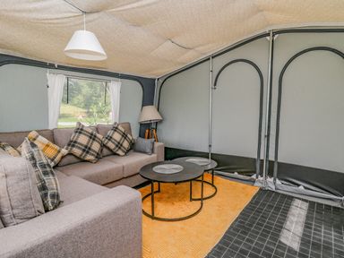 An indoor seating area with a sofa, plaid cushions, two round tables, a floor lamp, and a window with white curtains at The Heron in Nantmel near Rhayader