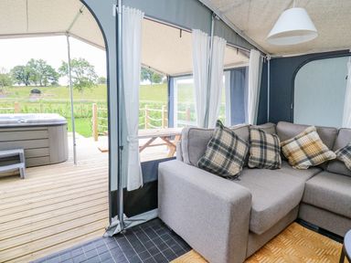 An indoor seating area with a gray couch and plaid pillows next to an outdoor wooden deck with a hot tub and picnic table at The Heron in Nantmel near Rhayader