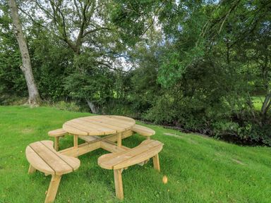 A wooden round picnic table with attached benches on grass near trees and a small stream at The Heron in Nantmel near Rhayader