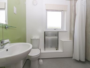 A bathroom with a white sink, toilet, window, heated towel rail, and a shower with a white curtain at 21 Eamont Park Penrith