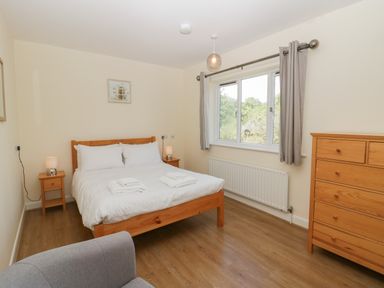 A bedroom with a double bed bedside tables a chest of drawers a window with curtains and a grey armchair at 12 Eamont Park in Penrith