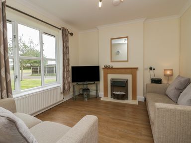 A living room with two beige sofas a television on a glass stand a fireplace and a window with curtains at 9 Eamont Park in Penrith