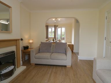 A living room with a sofa two cushions a side table with a lamp and a fireplace leading to a dining area at 9 Eamont Park in Penrith