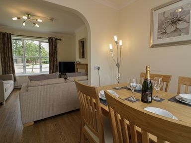 A dining table set with plates glasses and a bottle next to a living room with sofas a window and curtains at 9 Eamont Park in Penrith
