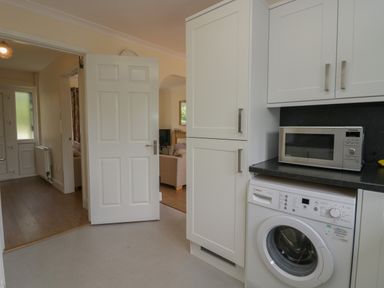A kitchen area with white cabinets a microwave and a washing machine and a door leading to a hallway and living room at 9 Eamont Park in Penrith