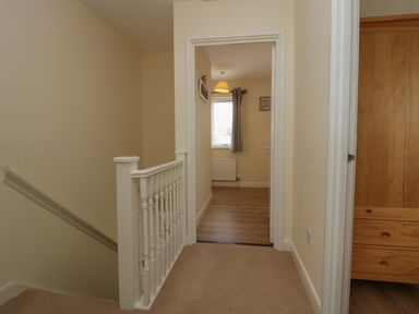 A hallway with carpeted floor and white railing leading to a room with wooden floor and a window at 9 Eamont Park in Penrith