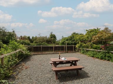 A picnic table on a gravel area with champagne bottle glasses and a newspaper surrounded by a wooden fence and greenery at 9 Eamont Park in Penrith