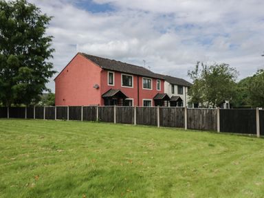 A row of houses with a red and white exterior behind a wooden fence and a grassy field with trees at 9 Eamont Park in Penrith