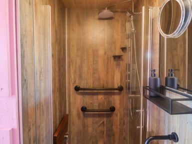 A shower area with wooden panel walls grab bars a rain showerhead and a small shelf with black soap dispensers at Sea Spray in Lyme Regis