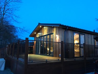 A wooden cabin with glass-railed deck and large windows at Loubi's Lakeside Lodge M1 Fallbarrow Park near Bowness-On-Windermere
