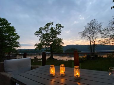 A patio with a wicker chair and table holding three lit candle jars overlooking a lake with trees at Loubi's Lakeside Lodge M1 Fallbarrow Park near Bowness-On-Windermere